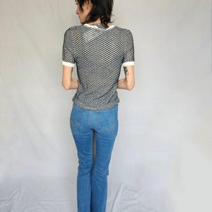 May include: A person wearing a black and white mesh shirt with a white trim and blue jeans.