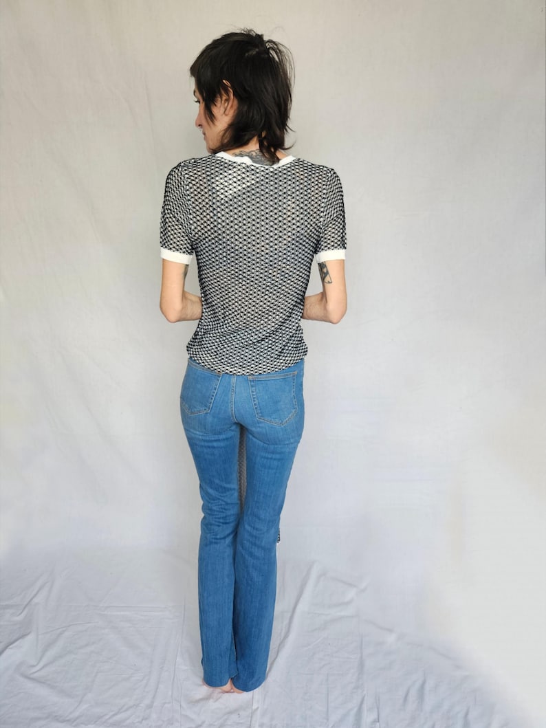 May include: A person wearing a black and white mesh shirt with a white trim and blue jeans.