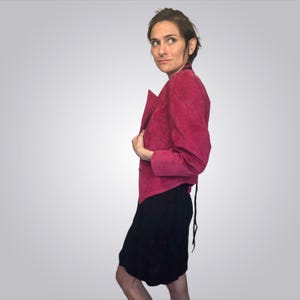 May include: A magenta suede jacket with a tailored silhouette. The jacket features a notched collar and button closure. The model is also wearing a black skirt. The jacket's color is a vibrant, saturated hue.