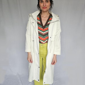 May include: A white long-sleeved hooded jacket with a drawstring waist and a zipper closure. The jacket is worn over a striped shirt and yellow pants.
