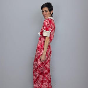 A woman wearing a red and white checkered dress stands against a gray background, looking over her shoulder.