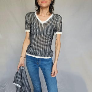 May include: A black and white patterned mesh top with a white v-neck and short sleeves. The top is worn with blue jeans.