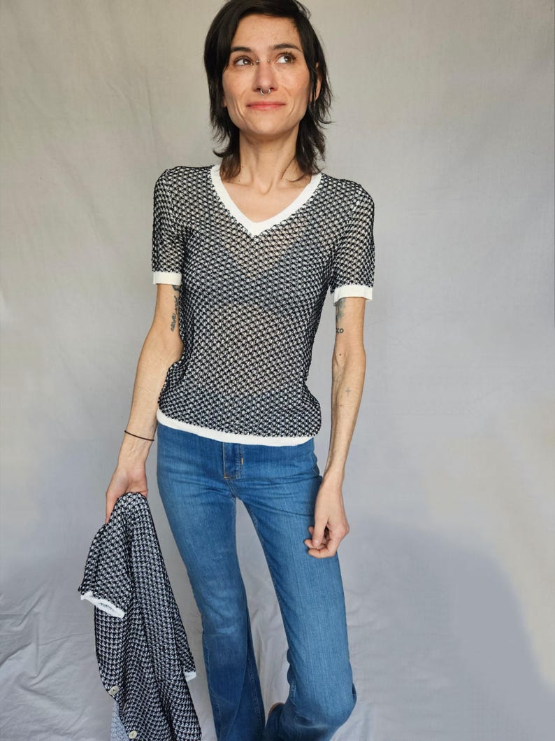 May include: A black and white patterned mesh top with a white v-neck and short sleeves. The top is worn with blue jeans.