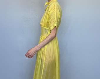 May include: A sheer, bright yellow dress with short sleeves and a high collar. The dress has a fitted waist and a flowing skirt. The fabric appears lightweight and slightly translucent. The model is wearing the dress.