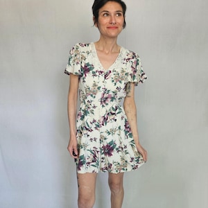 Puede incluir: Un mono crema con estampado floral en tonos morados, verdes y marrones. Tiene un escote en V con ribete de encaje y mangas cortas con volantes. La tela parece ligera, ideal para el clima cálido.