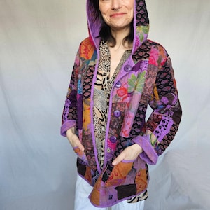 May include: A hooded patchwork jacket in a variety of colours and patterns. The jacket has a purple hood and trim, with a button-up front and pockets. The design incorporates floral, geometric, and animal print fabrics. The jacket is a unique piece of clothing.