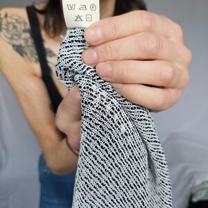 May include: A white tag with black text and symbols that reads "Made in Italy". The tag has a washing instruction symbol for machine wash, a symbol for tumble dry low, a symbol for iron low, and a symbol for dry clean. The tag is being held by a person's hand.