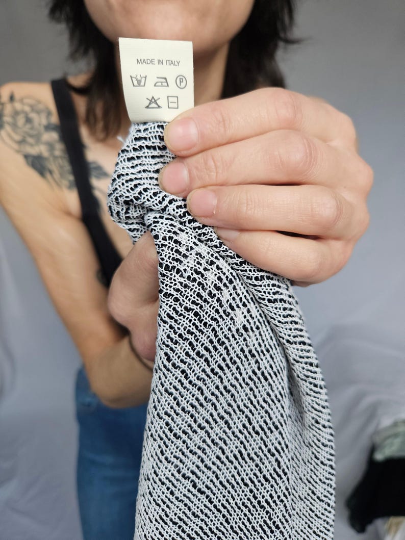 May include: A white tag with black text and symbols that reads "Made in Italy". The tag has a washing instruction symbol for machine wash, a symbol for tumble dry low, a symbol for iron low, and a symbol for dry clean. The tag is being held by a person's hand.