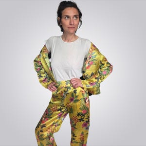 May include: A white crew neck t-shirt paired with a matching yellow blazer and trousers set. The blazer and trousers feature a vibrant floral pattern with green leaves, pink flowers, and small portraits of Frida Kahlo printed throughout the fabric.