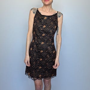 May include: Black lace dress with a floral pattern in gold. The dress features a scoop neckline, spaghetti straps, and a scalloped hem. The knee-length dress is a classic design.