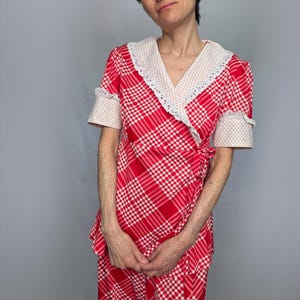 A woman wearing a red and white checkered dress with a lace collar and sleeves stands against a gray background.