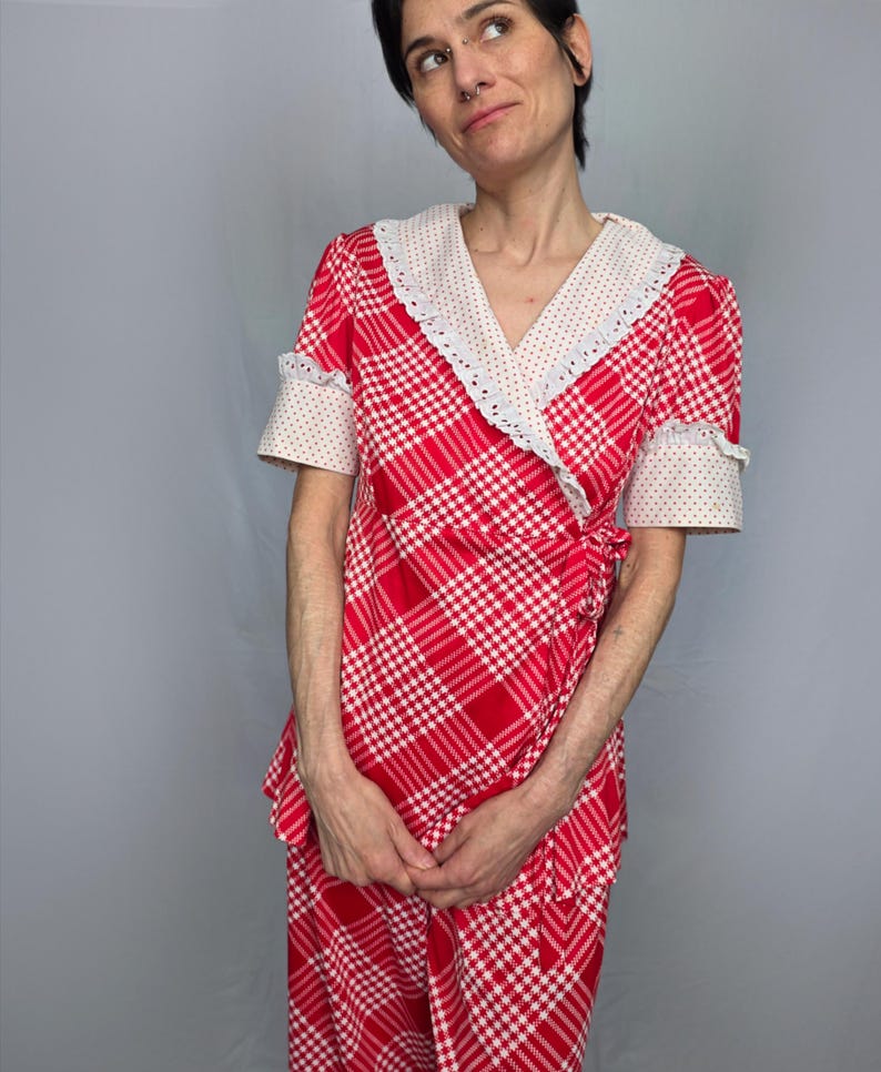 A woman wearing a red and white checkered dress with a lace collar and sleeves stands against a gray background.
