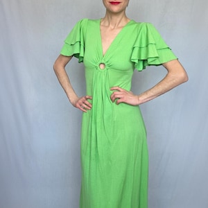 May include: A vibrant green maxi dress with a V-neck and a ring detail at the waist. The dress has ruffled sleeves and a flowing skirt. The model is wearing the dress.