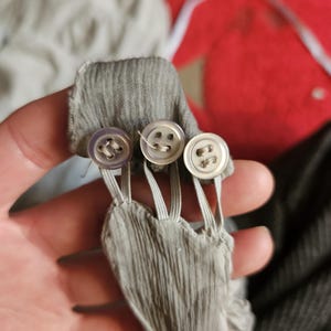 May include: Close-up of a gray fabric detail with three small, round buttons. The buttons are attached to the fabric with elastic loops. The fabric has a textured, pleated appearance.