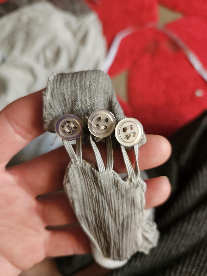 May include: Close-up of a gray fabric detail with three small, round buttons. The buttons are attached to the fabric with elastic loops. The fabric has a textured, pleated appearance.