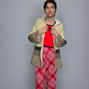 A woman wearing a beige jacket, red top, and red and white plaid pants stands against a gray background, looking to her left with a neutral expression.