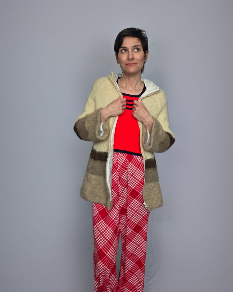 A woman wearing a beige jacket, red top, and red and white plaid pants stands against a gray background, looking to her left with a neutral expression.