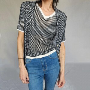 May include: A black and white patterned mesh shirt layered over a white v-neck shirt. The person is wearing blue jeans.