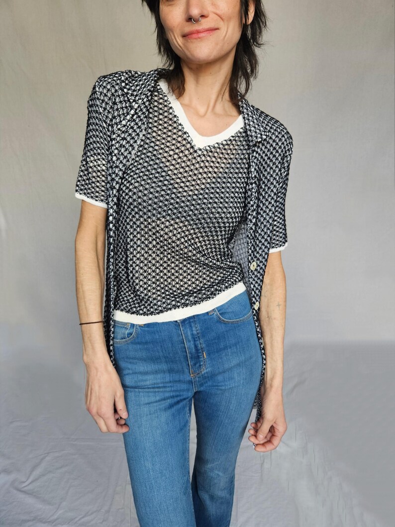 May include: A black and white patterned mesh shirt layered over a white v-neck shirt. The person is wearing blue jeans.