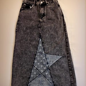 Può includere: Gonna lunga in denim con lavaggio acido, a vita alta e con spacco frontale. La gonna presenta un ampio pannello a forma di stella realizzato in denim più chiaro con un motivo a stelle. Design classico a cinque tasche.