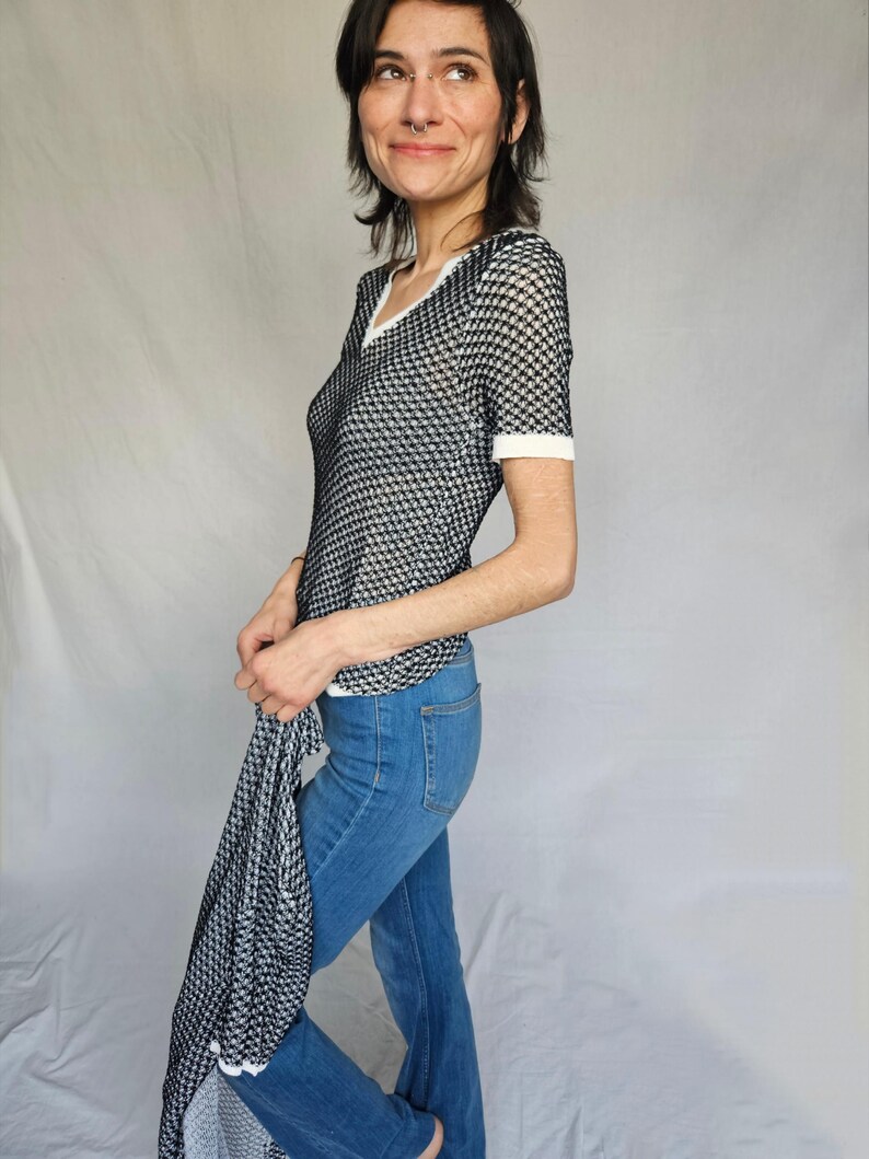 May include: A black and white patterned mesh top with a white trim around the neckline and short sleeves. The top is paired with blue jeans.