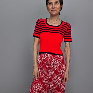 A woman wearing a red and black striped top and red and white plaid pants stands against a gray background, looking to her left with a neutral expression.