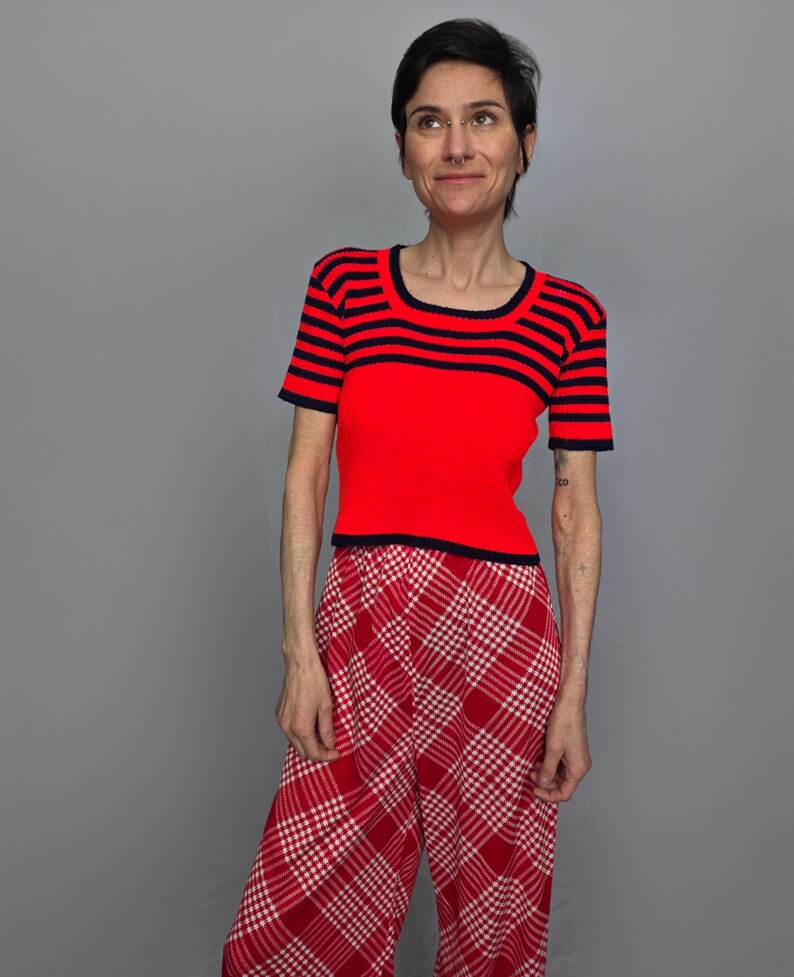 A woman wearing a red and black striped top and red and white plaid pants stands against a gray background, looking to her left with a neutral expression.
