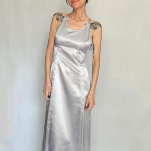 Puede incluir: Un vestido largo plateado de satén con cuello halter. El vestido tiene un corpiño ajustado y una falda fluida. La tela tiene un brillo sutil, que refleja la luz. El vestido es de estilo formal, adecuado para ocasiones especiales.
