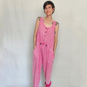 May include: A pink jumpsuit with a button-up front, drawstring waist, and pockets. The cuffs are rolled up, showing a darker pink lining. The sleeveless top and loose-fitting trousers create a casual, comfortable style.