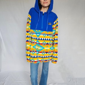 May include: A blue and yellow fleece pullover with a geometric pattern. The pullover has a hood and a zip closure. The zip is blue and has a small logo on the zip pull.