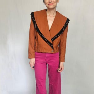 May include: A rust-coloured jacket with a wide collar and black trim. The jacket has a unique design with a wrap-around style and button closure. Paired with bright pink trousers.