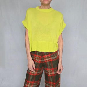 Puede incluir: Una persona viste un top amarillo neón de manga corta y pantalones a cuadros. Los pantalones tienen un patrón de cuadros rojos, marrones y beige. La parte superior es de color liso y brillante. El atuendo es casual y elegante.