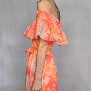 A woman wearing a vibrant, floral-patterned dress with off-the-shoulder sleeves stands against a plain background.