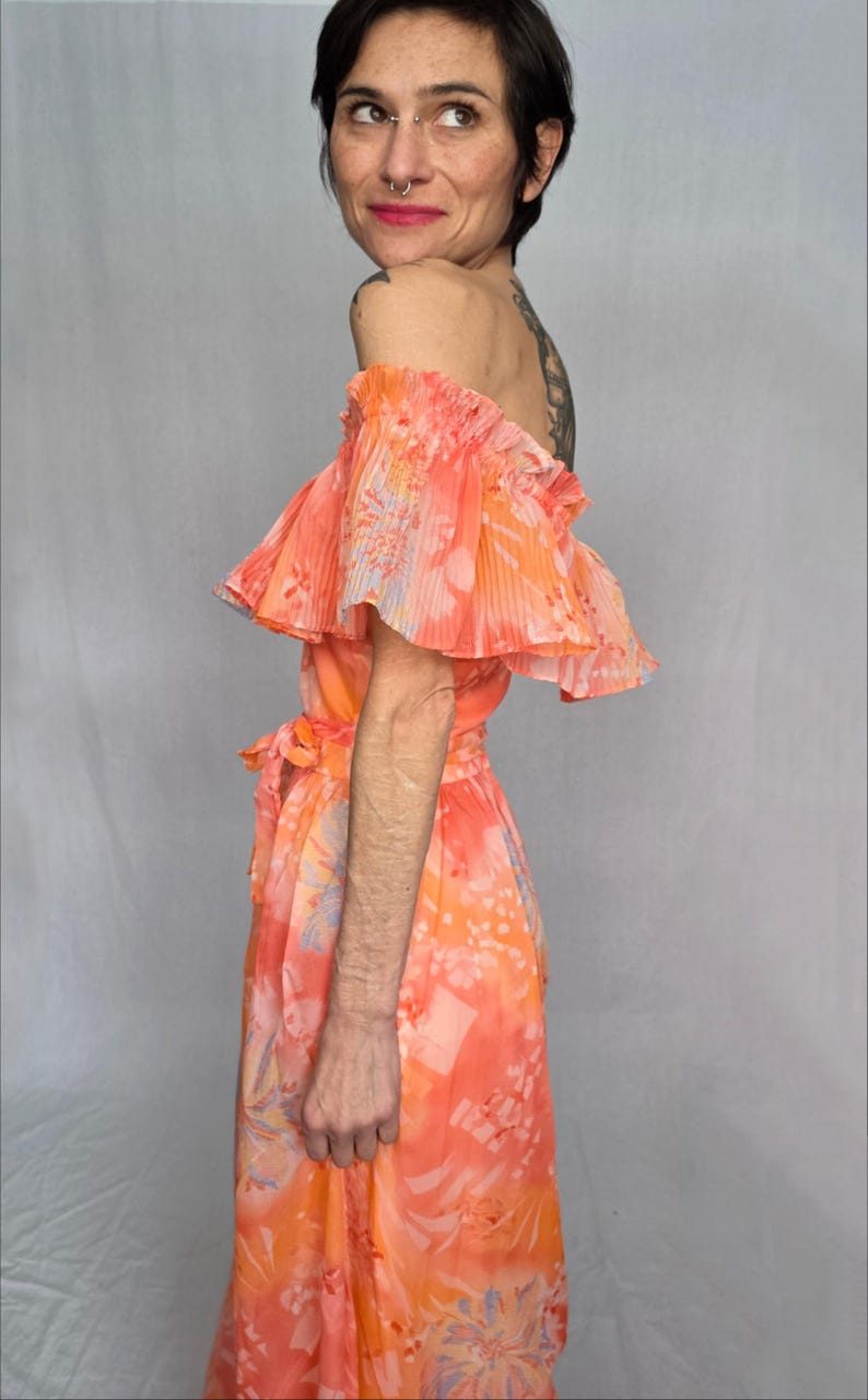 A woman wearing a vibrant, floral-patterned dress with off-the-shoulder sleeves stands against a plain background.