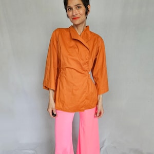 May include: An orange wrap top with three-quarter sleeves and a single button closure. Paired with bright pink, wide-leg pants. The outfit is styled on a person in front of a neutral background.