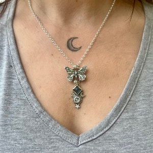May include: A silver necklace with a butterfly charm, a moonstone pendant, and a crescent moon charm. The necklace is on a person's chest.