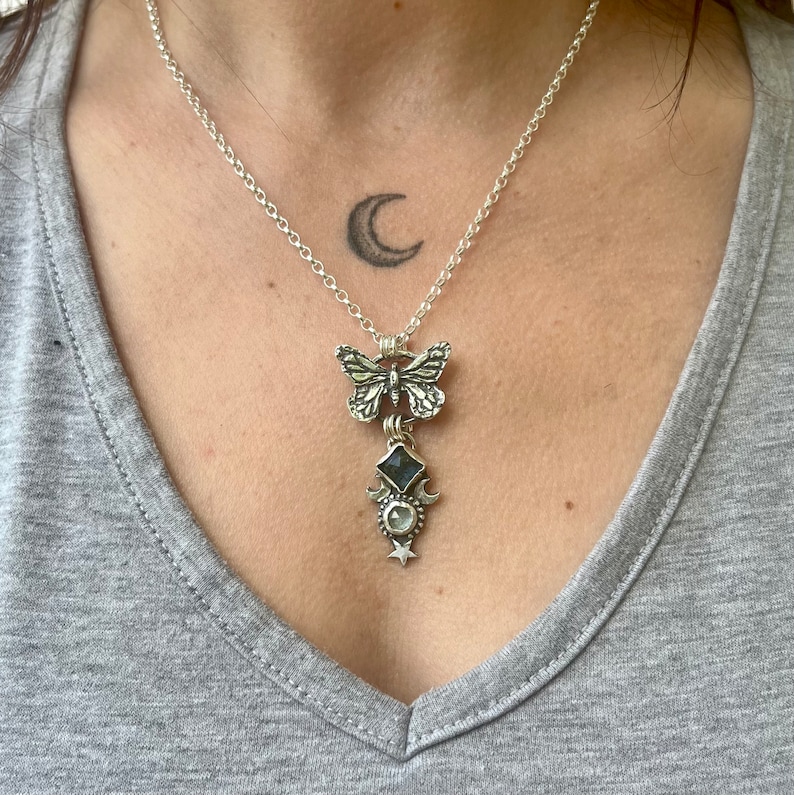 May include: A silver necklace with a butterfly charm, a moonstone pendant, and a crescent moon charm. The necklace is on a person's chest.
