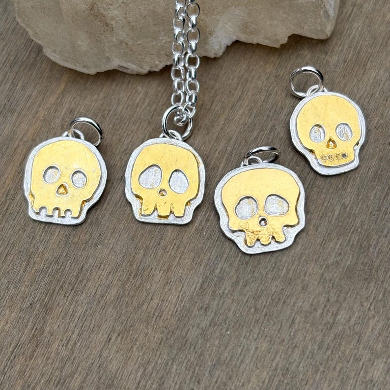Op de afbeelding: Zilver- en goudkleurige schedelhangers. De hangers hebben een gouden schedelontwerp met zilveren randen. E&eacute;n hanger hangt aan een zilveren ketting. De schedels hebben oog- en neusgaten en een grillige kaaklijn.
