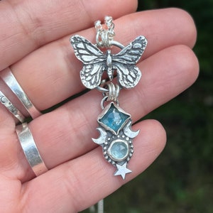 May include: A silver necklace with a butterfly charm and a pendant featuring two blue gemstones, crescent moons, and a star.