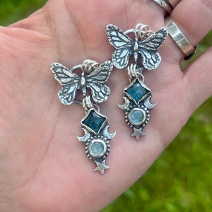 May include: Silver butterfly earrings with blue gemstone accents. The earrings feature a crescent moon and star design.