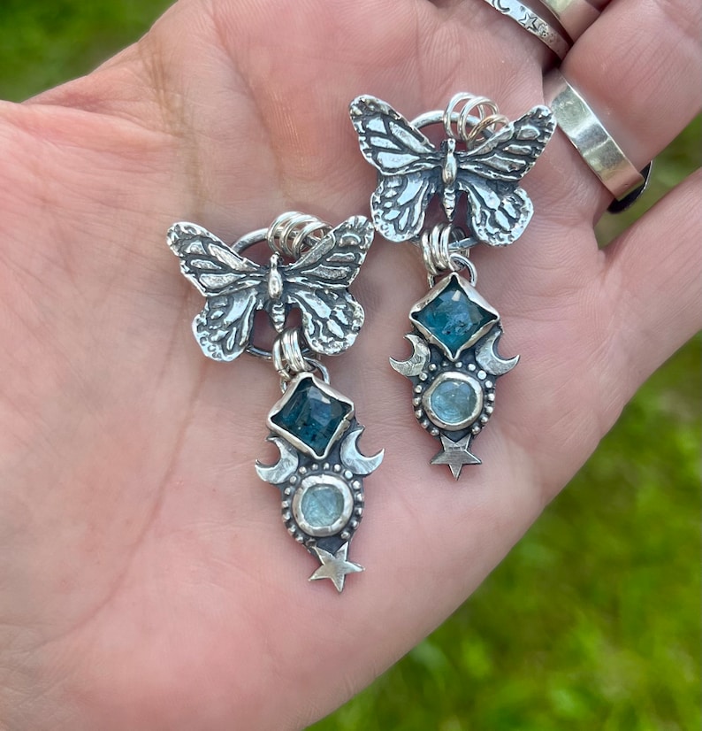 May include: Silver butterfly earrings with blue gemstone accents. The earrings feature a crescent moon and star design.