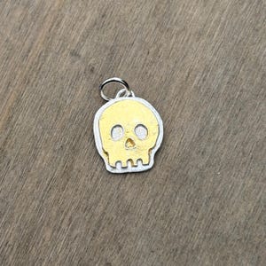 Op de afbeelding: Zilverkleurige schedel hanger met een goudkleurig schedel ontwerp. De hanger heeft een kleine lus aan de bovenkant om aan een ketting of armband te bevestigen. De schedel heeft twee oogkassen en een neusholte.