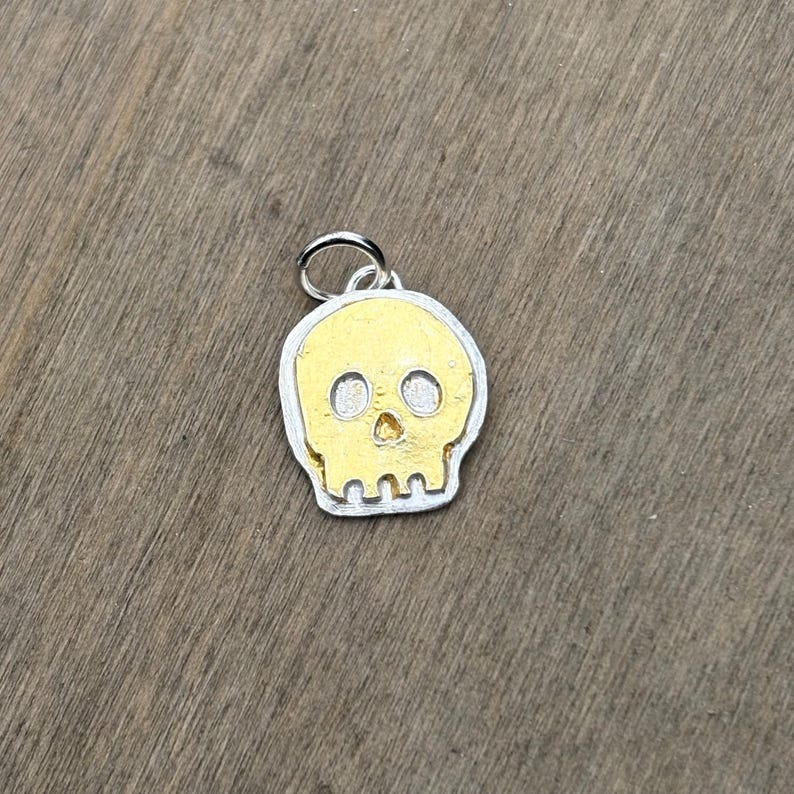 Op de afbeelding: Zilverkleurige schedel hanger met een goudkleurig schedel ontwerp. De hanger heeft een kleine lus aan de bovenkant om aan een ketting of armband te bevestigen. De schedel heeft twee oogkassen en een neusholte.