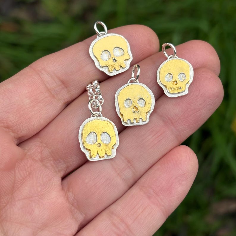 Op de afbeelding: Vier kleine schedelhangers met een zilveren rand en een goudkleurig schedelontwerp. E&eacute;n hanger is bevestigd aan een zilveren ketting. De hangers worden in een hand gehouden tegen een vage groene achtergrond.