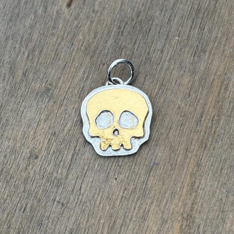 Op de afbeelding: Een zilver- en goudkleurige schedel hanger. De schedel heeft een goudkleurig gezicht met zilverkleurige contouren en een zilveren lus voor een ketting. De hanger ligt op een houten oppervlak.