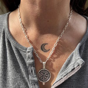 May include: A silver chain necklace with a circular pendant featuring a compass design and runes. The pendant is hanging on a person's chest. A crescent moon tattoo is visible on the person's chest.