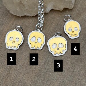 Op de afbeelding: Vier zilver-gouden schedelhangers. Elke hanger heeft een gouden schedelontwerp met zilveren contouren. De hangers zijn genummerd van 1 tot 4. E&eacute;n hanger is bevestigd aan een zilveren ketting.