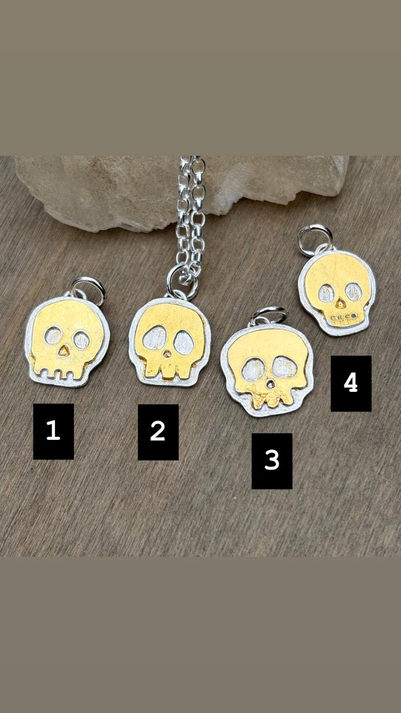 Op de afbeelding: Vier zilver-gouden schedelhangers. Elke hanger heeft een gouden schedelontwerp met zilveren contouren. De hangers zijn genummerd van 1 tot 4. E&eacute;n hanger is bevestigd aan een zilveren ketting.