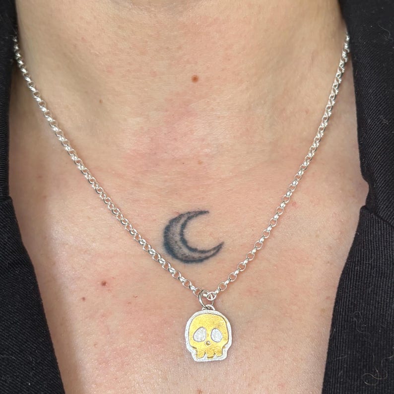 Op de afbeelding: Zilveren ketting met een kleine schedel hanger. De schedel hanger heeft een goudkleurig gezicht en een zilveren omtrek. De ketting wordt om de nek gedragen.