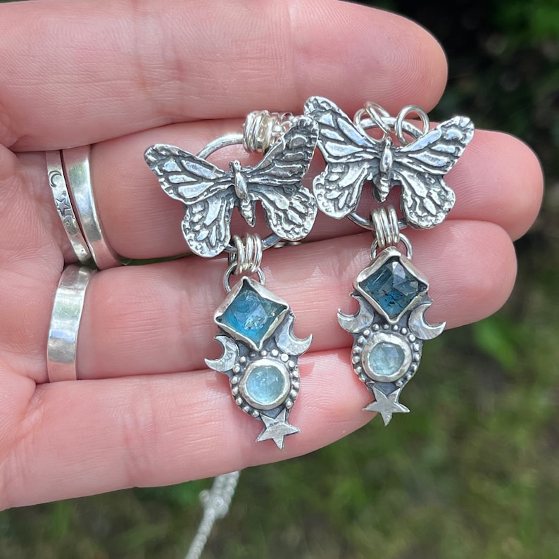 May include: Silver butterfly earrings with blue and green gemstone accents. The earrings feature a crescent moon and star design.